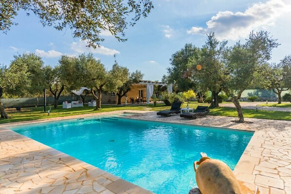 Une piscine entourée d’arbres, avec des chaises longues et une pergola, ainsi qu’une terrasse en pierre.