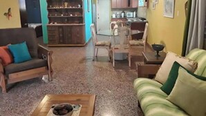 A living room with a sofa, chairs, and a coffee table. There is a kitchen area with cabinets and a refrigerator in the background.