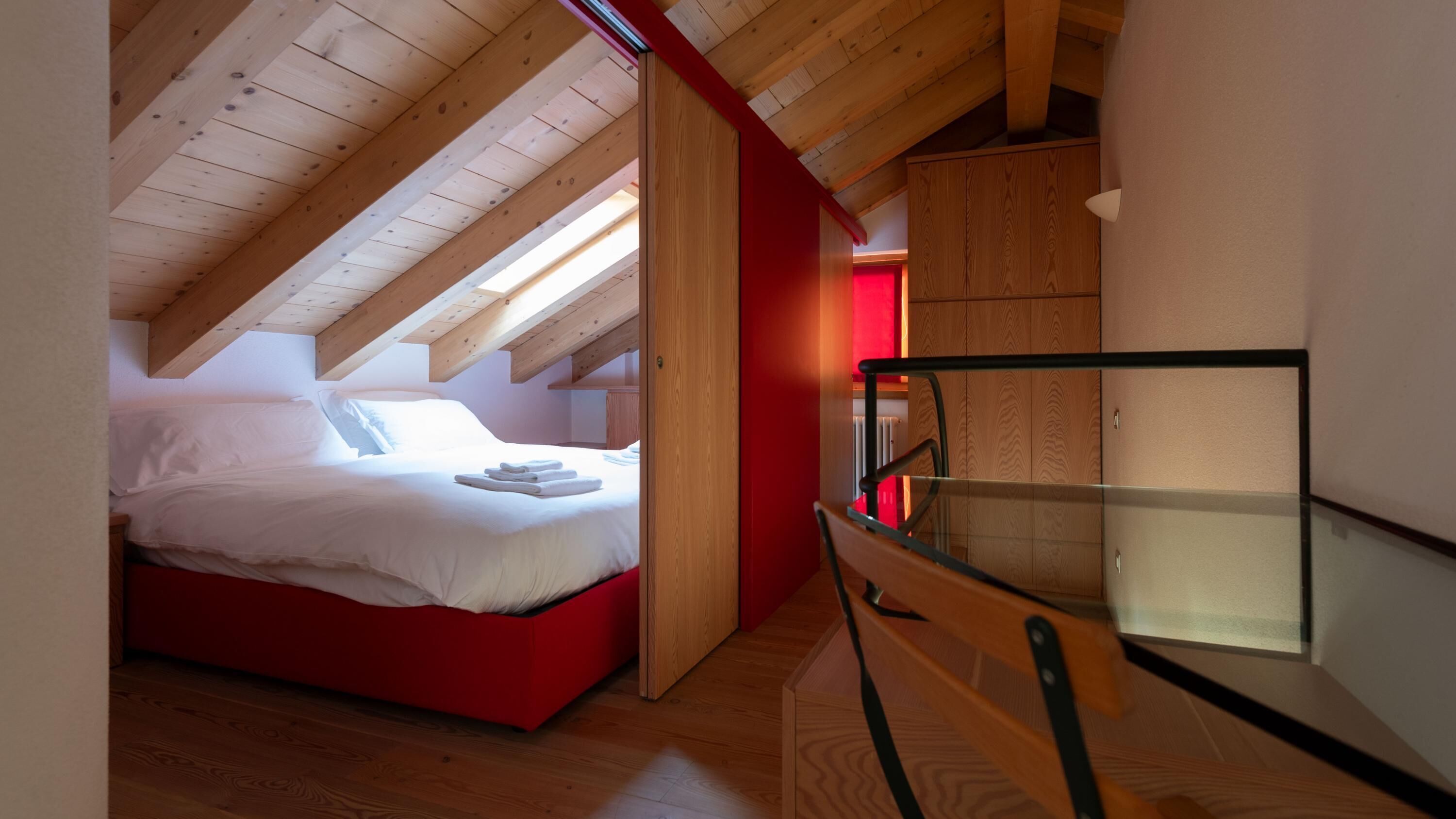 A bedroom with a sloped ceiling, a bed, a red accent wall, and a glass railing.
