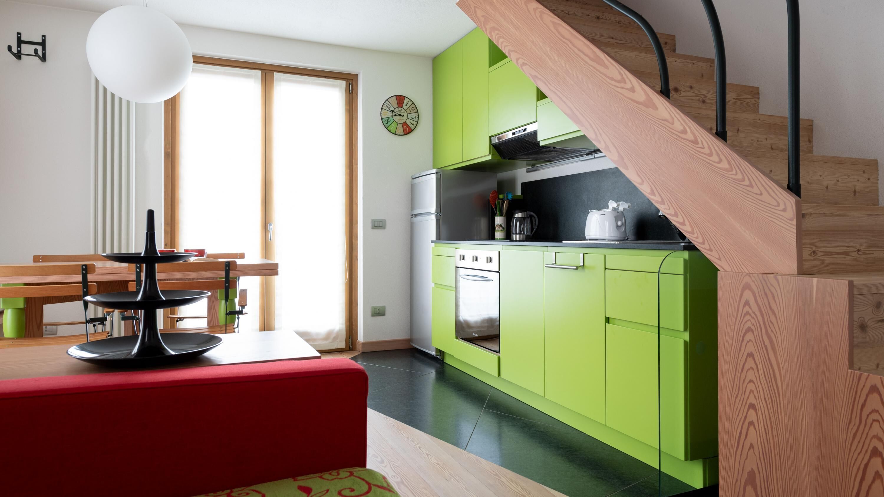 A modern kitchen with green cabinets, a wooden staircase, and a dining area with a red bench.