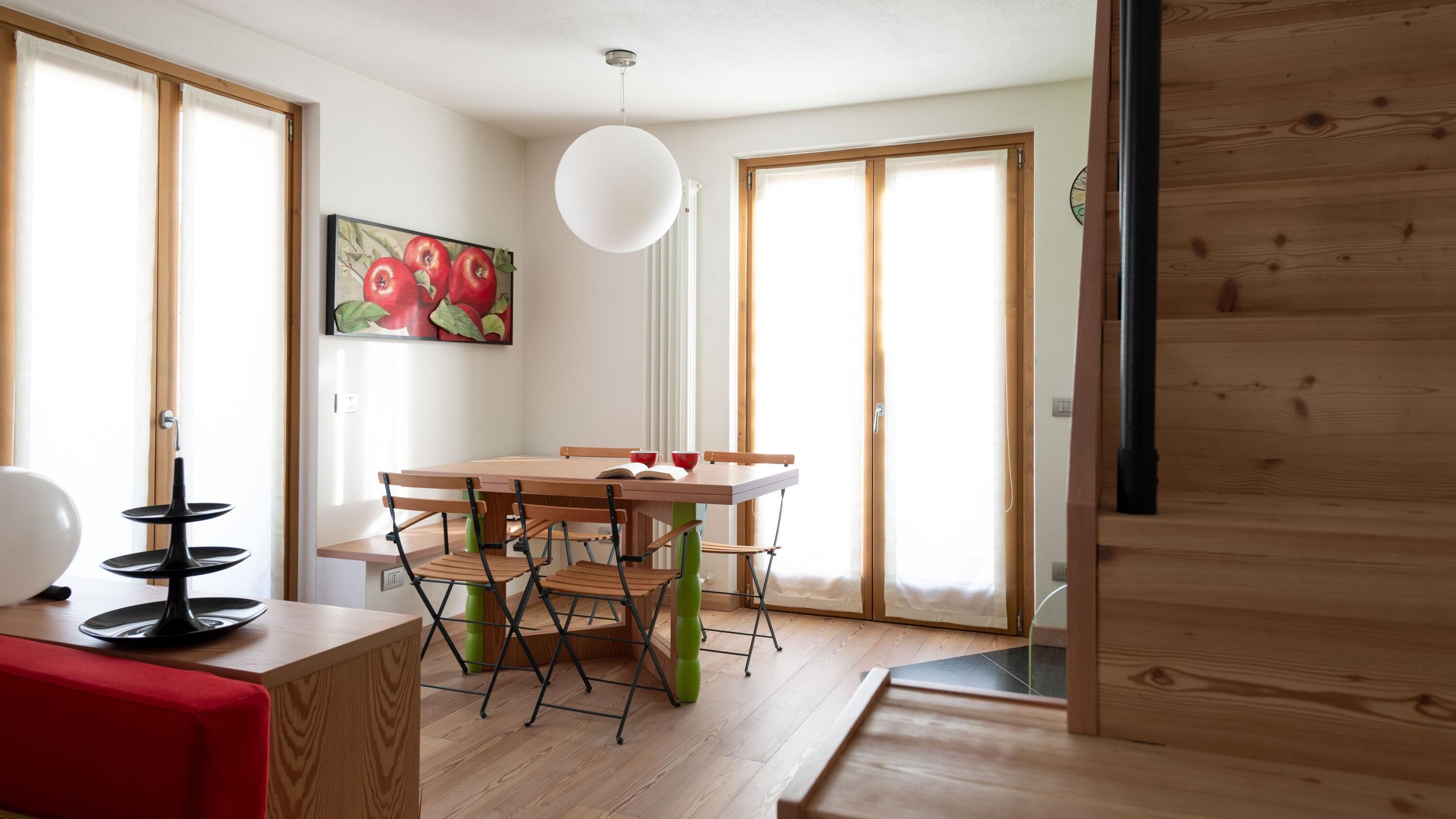A dining area with a wooden table and chairs, a painting of apples, and a large window with curtains.