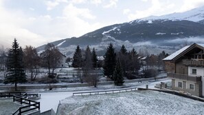 Paisatge cobert de neu amb una muntanya al fons, un camí i un edifici amb un balcó.