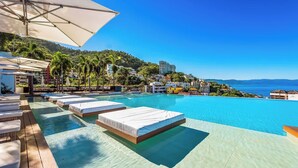 Zona de piscina con sillones de descanso y una cama, con vista a un mar azul cristalino y una colina con edificios.
