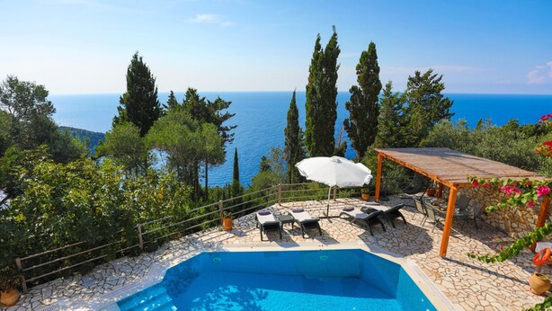 Une piscine entourée d’une terrasse en pierre, de chaises longues et d’une pergola avec un parasol.