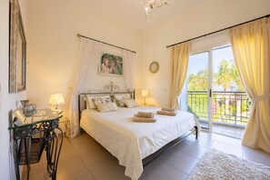 Quarto com uma cama grande, uma mesa de vidro com abajur, um quadro na parede e uma varanda com cortinas.