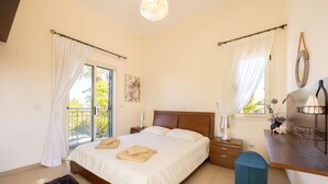 A bedroom with a bed, bedside tables, a desk, a chair, a window with curtains, and a view of greenery outside.