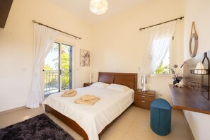 A bedroom with a bed, bedside tables, a desk, a chair, a window with curtains, and a view of greenery outside. - Latchi Aria Villa in Latchi (Neo Chorio)