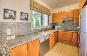 A kitchen with granite countertops, wooden cabinets, a stainless steel refrigerator, and a window with a view of greenery.