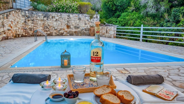 A poolside setting with a table set for a meal, including bread, olives, and a bottle of ouzo. - Beach House Sophia in Paxi (Paxos)