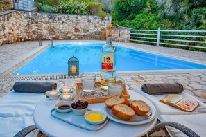 A poolside setting with a table set for a meal, including bread, olives, and a bottle of ouzo. - Beach House Sophia in Paxi (Paxos)