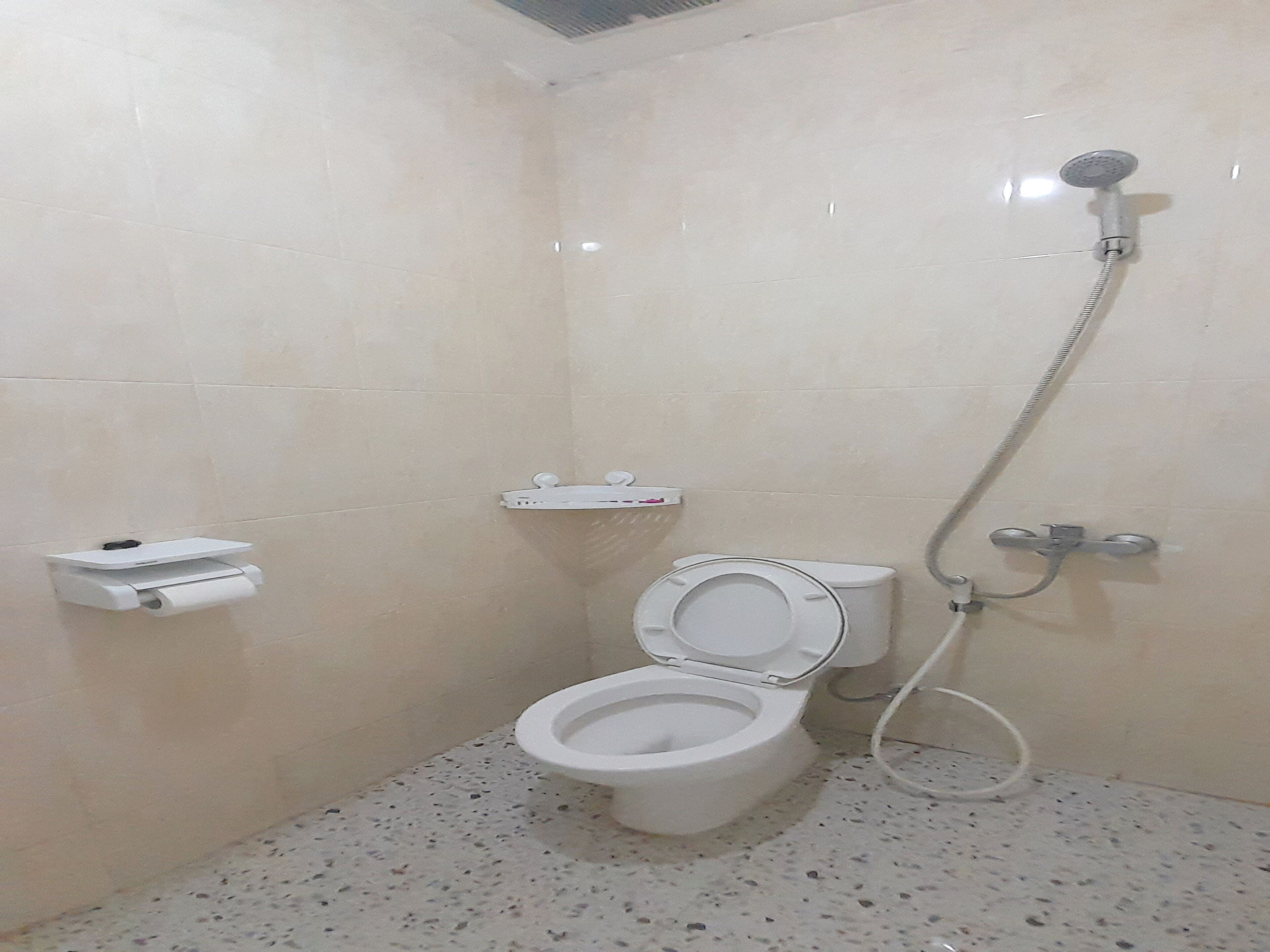 Une petite salle de bain carrelée, équipée de toilettes, d’une douche et d’un distributeur d’essuie-tout.