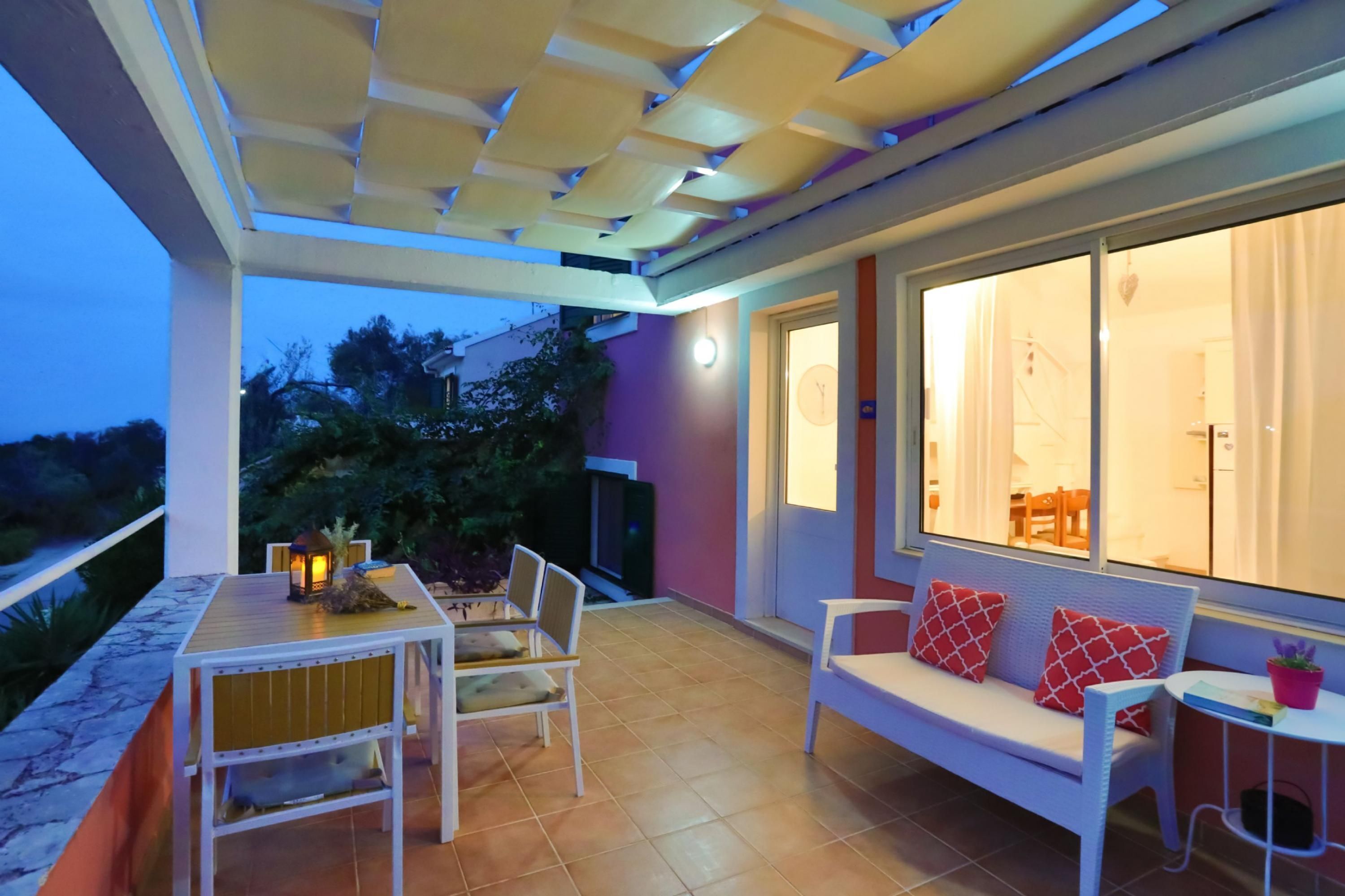 Un espace extérieur aménagé en terrasse, avec une table et des chaises, un canapé blanc agrémenté de coussins rouges et une grande fenêtre donnant sur l’intérieur.