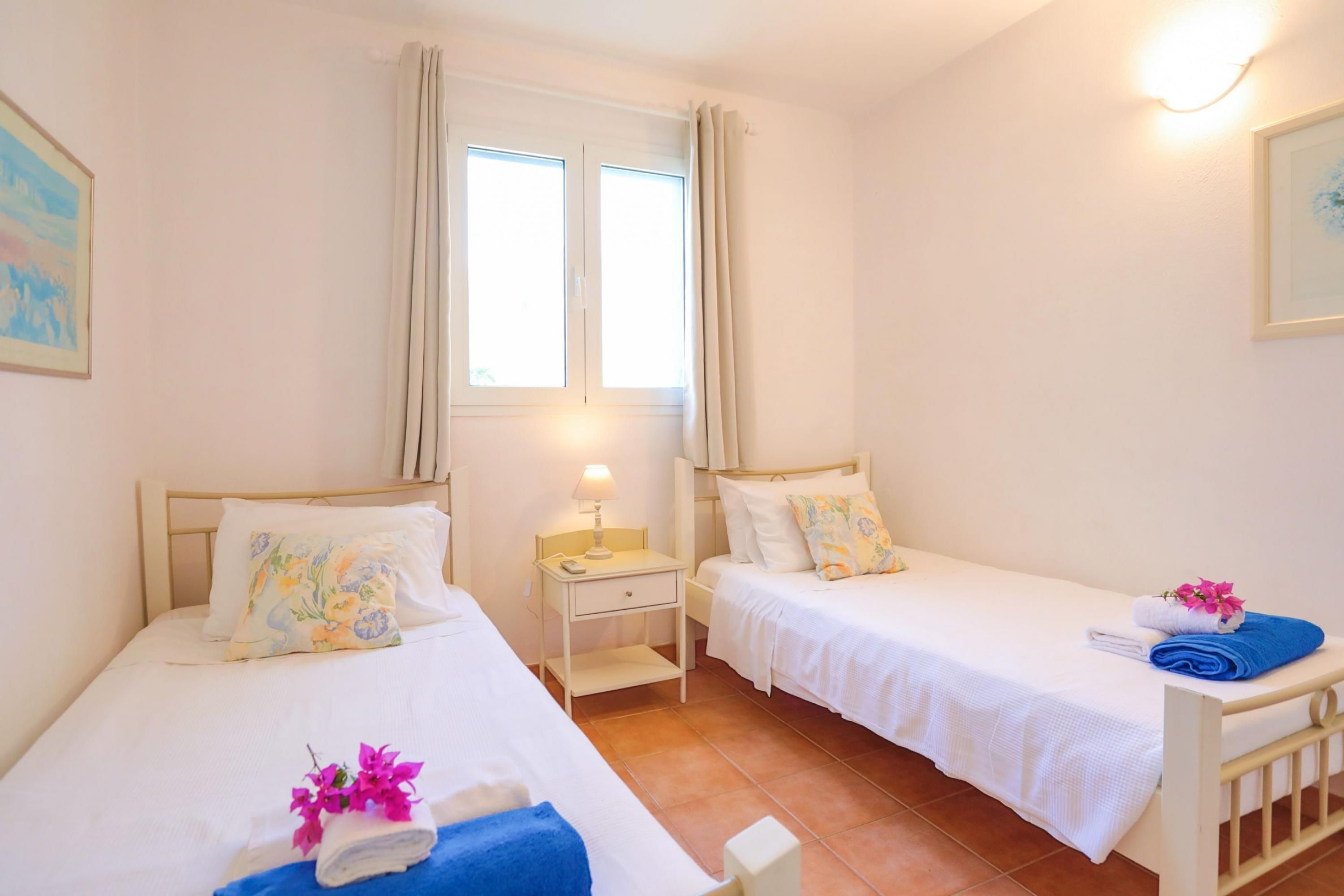 A hotel room with two single beds, a window with curtains, a bedside table with a lamp, and a wall-mounted light fixture.