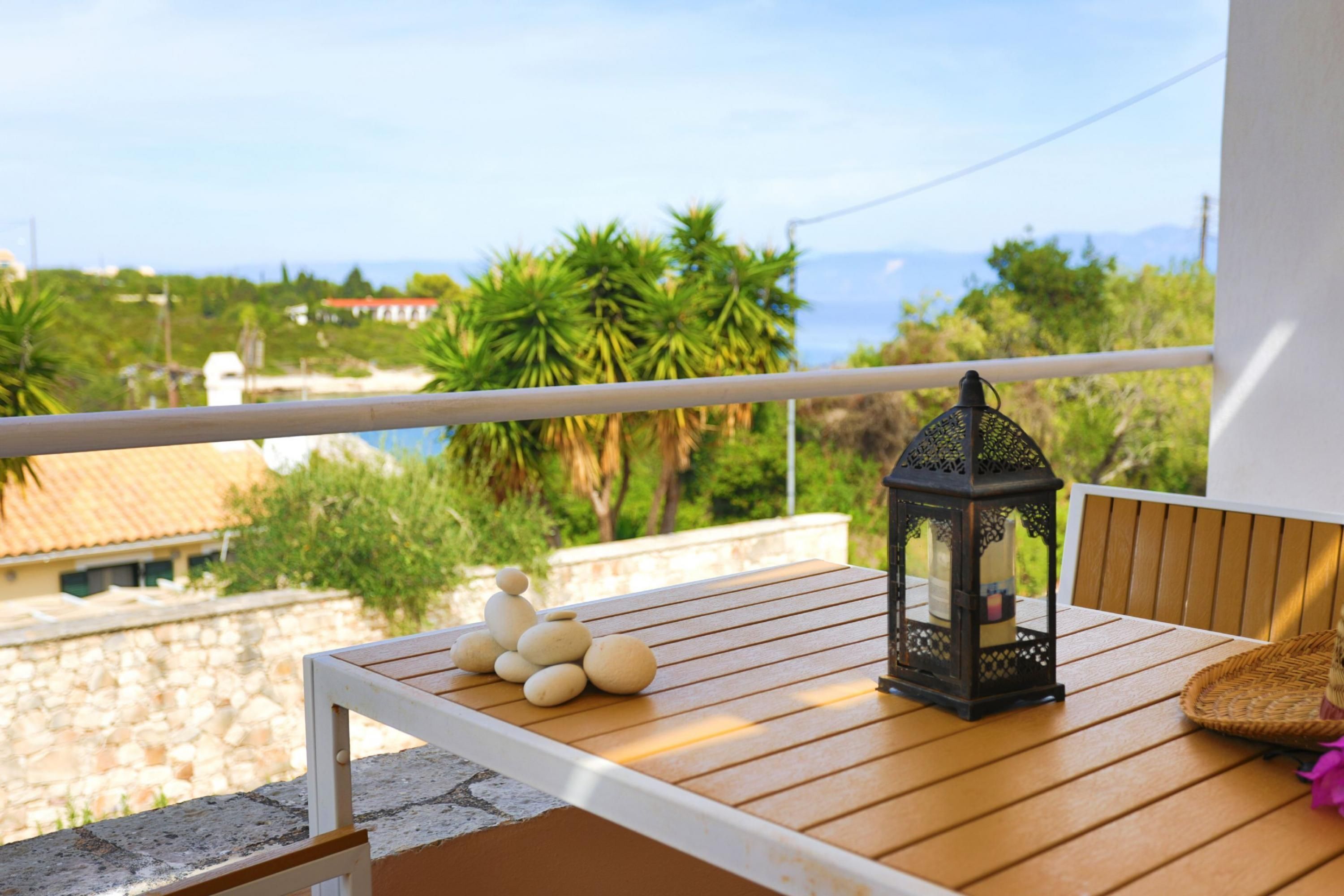 Una mesa de madera con un farol y piedras apiladas en un balcón con vistas a una zona costera con palmeras y edificios.