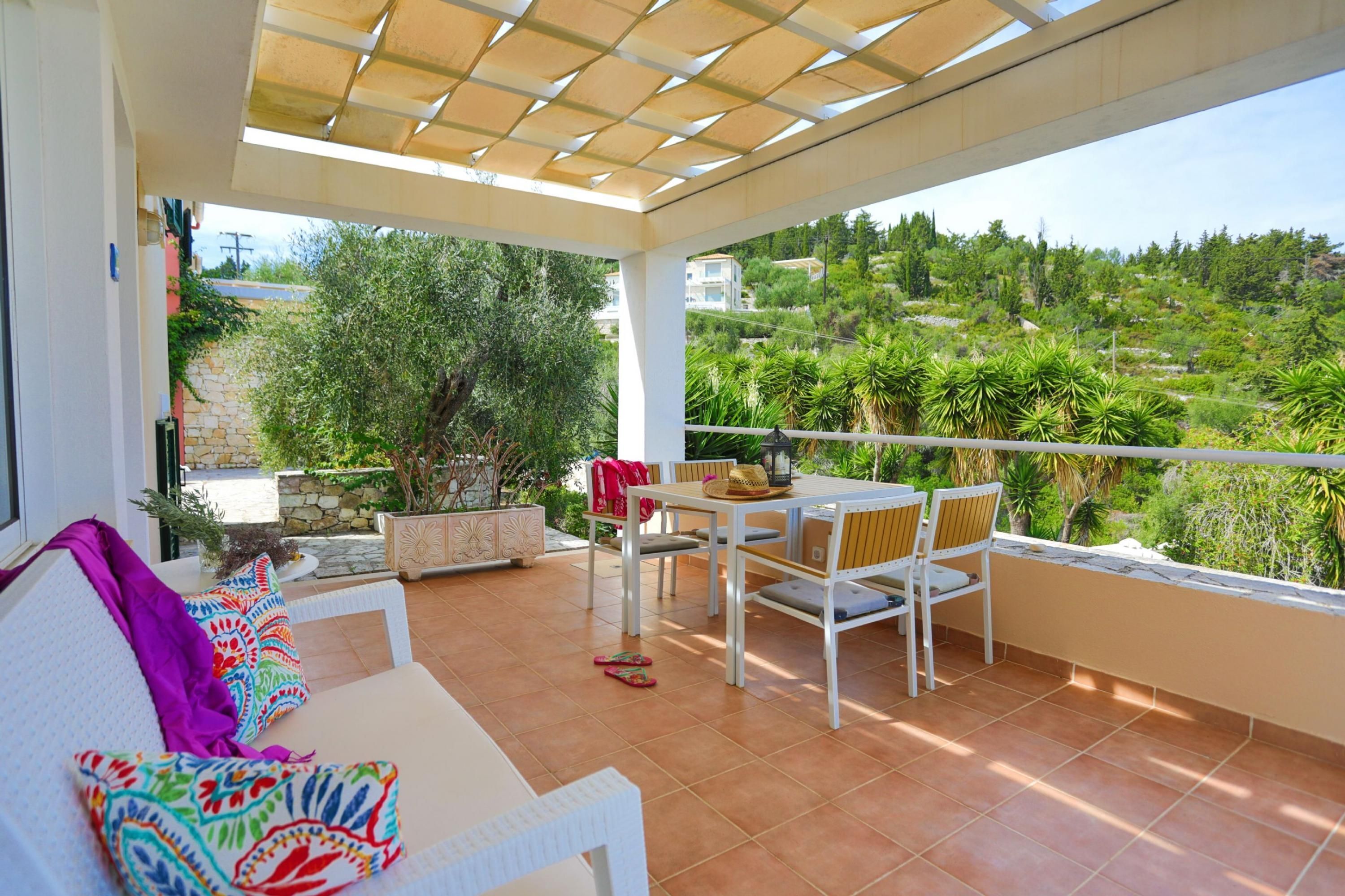 Une terrasse couverte avec une table et des chaises blanches, un canapé agrémenté de coussins colorés et une vue sur un paysage verdoyant.