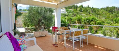 Une terrasse couverte avec une table et des chaises blanches, un canapé agrémenté de coussins colorés et une vue sur un paysage verdoyant.