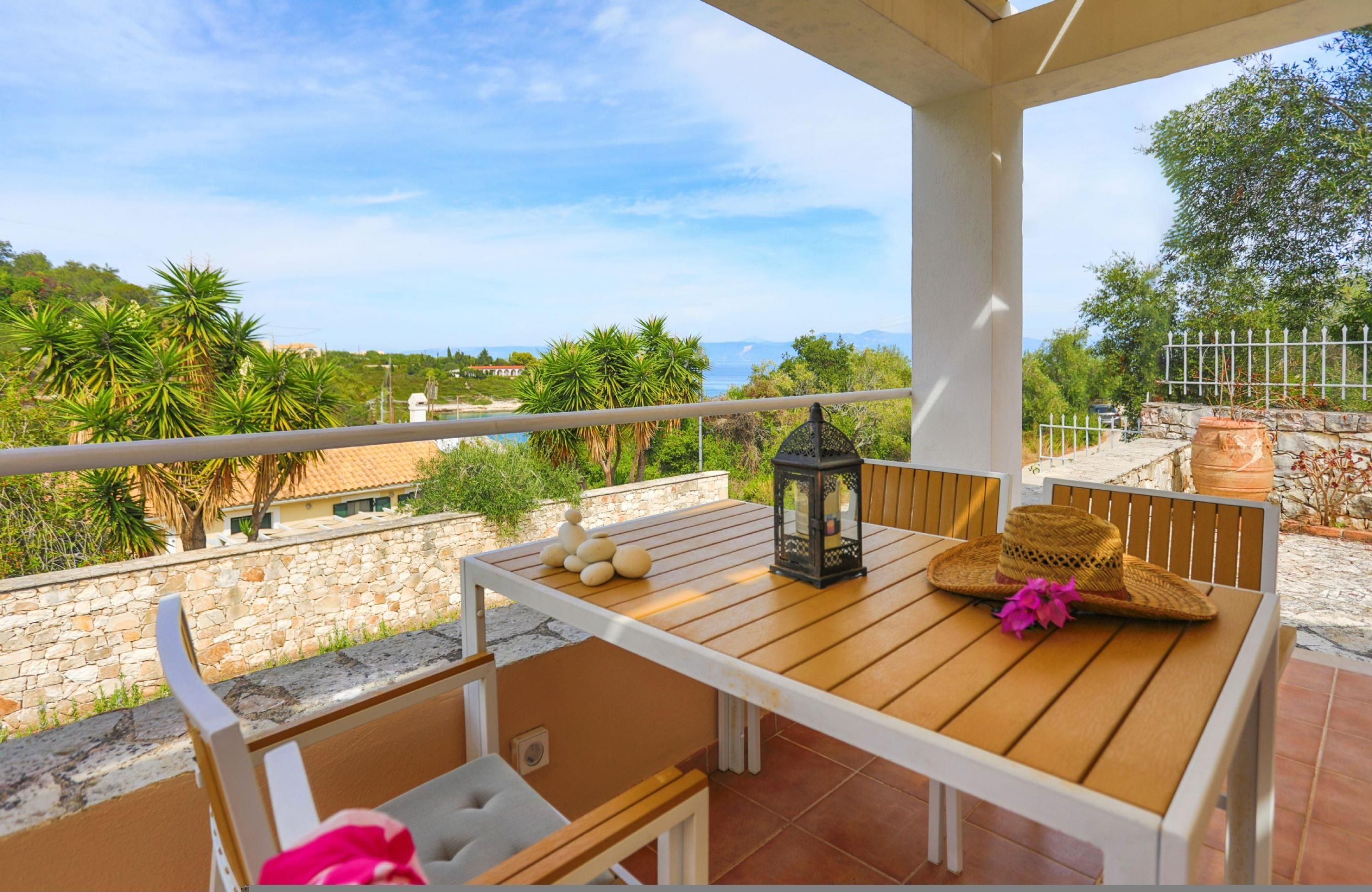A wooden table with a lantern and a straw hat on a balcony overlooking a scenic view with trees and a stone wall.