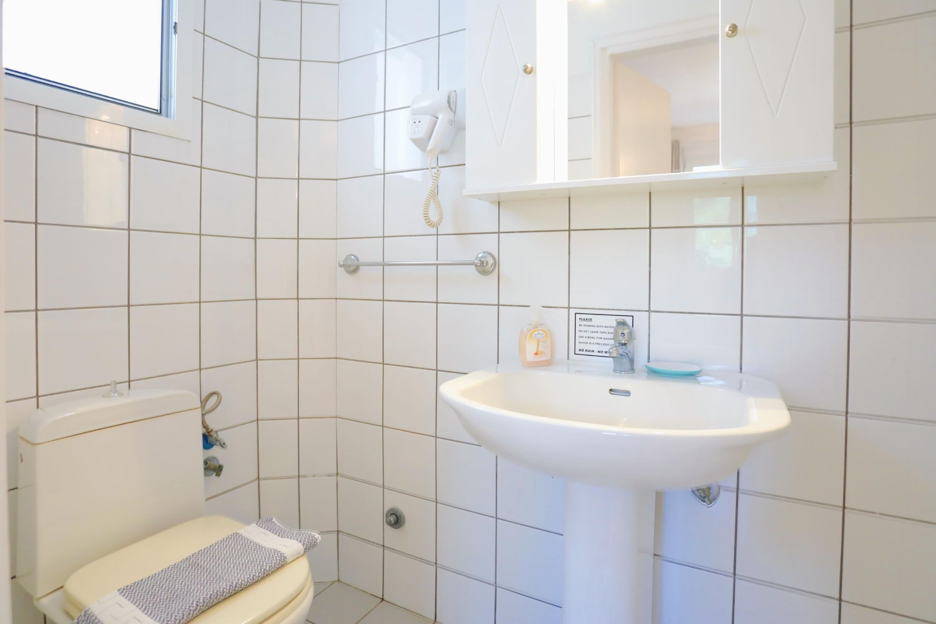 A bathroom with white tiles, a toilet, a sink, a mirror, and a hairdryer on the wall.