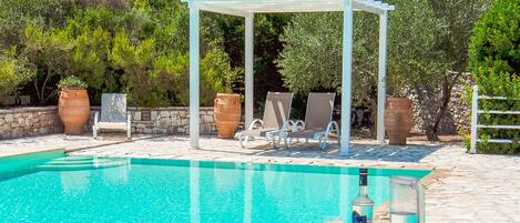 A pool area with a pergola, lounge chairs, and a table with fruits and drinks.