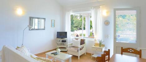 Una habitación luminosa y bien iluminada con un sofá, una mesita, un televisor y un ventanal con vistas a zonas verdes.