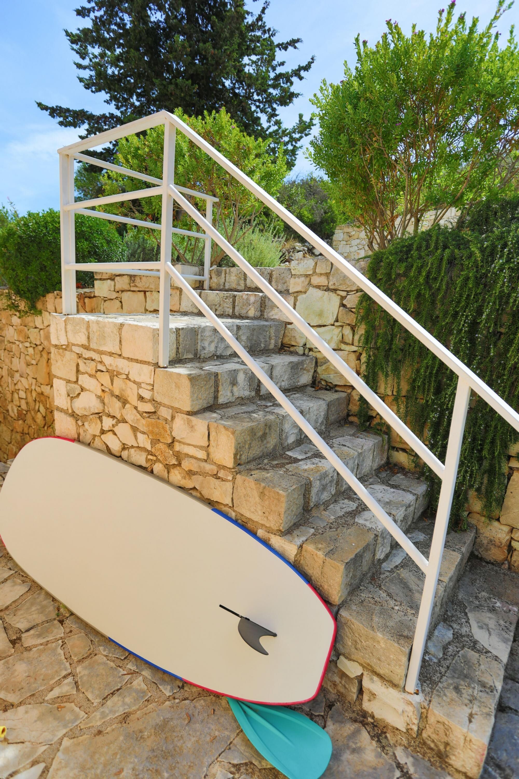 A stone staircase with a white railing, a surfboard leaning against it, and greenery in the background.