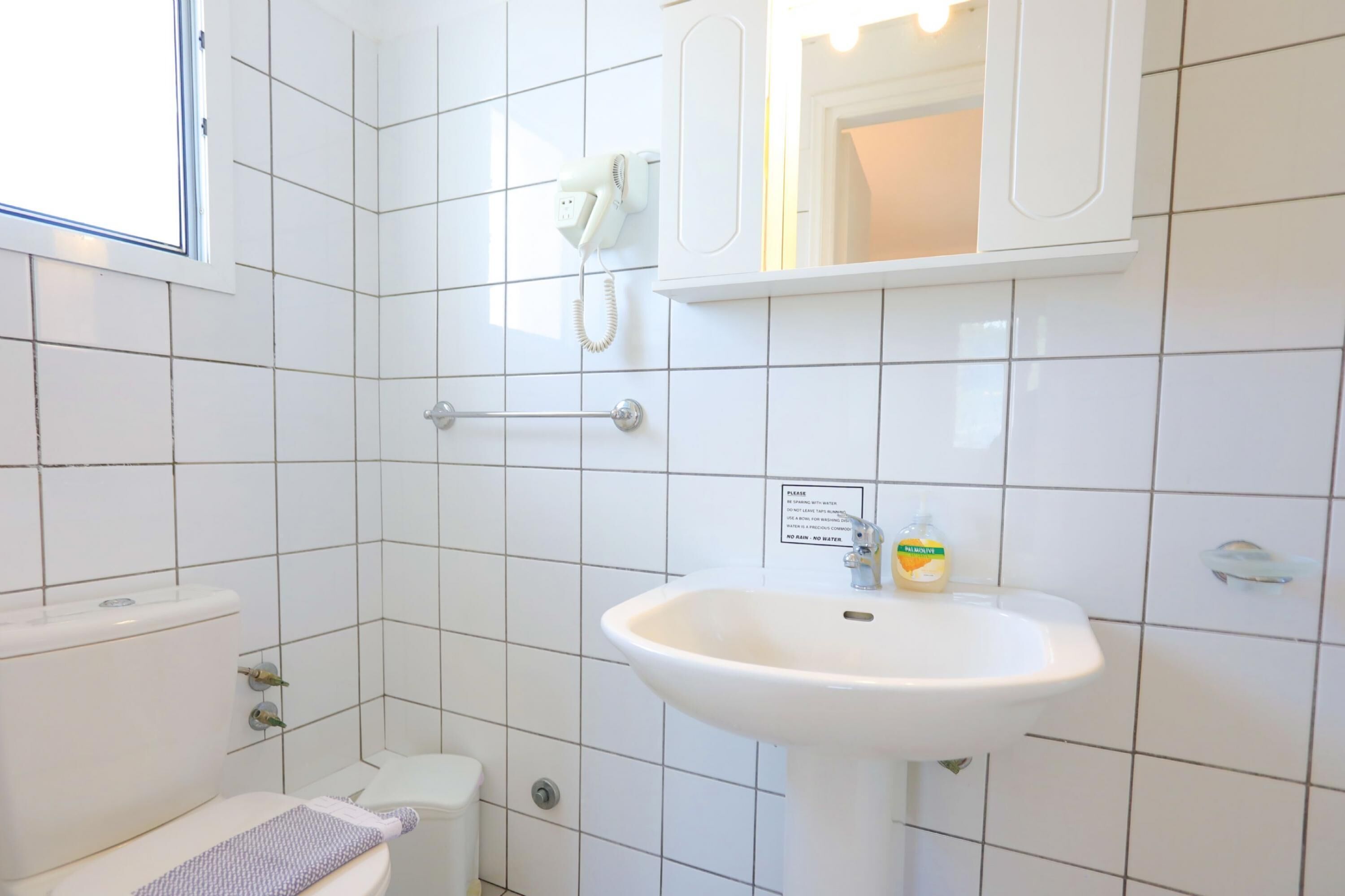 A bathroom with white tiles, a toilet, a sink with a soap dispenser, a hand dryer, and a mirror.