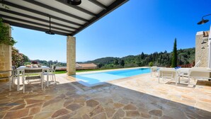 A covered outdoor swimming pool with stone flooring, white furniture, and a view of a mountainous landscape. - Lithari Tria in Agios Georgios (Corfu)