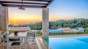 A poolside area with stone pillars, a table with chairs, and a view of a sunset over a hilly landscape. - Lithari Tria in Agios Georgios (Corfu)
