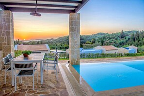 Ein Poolbereich mit Steinpfeilern, einem Tisch mit Stühlen und Blick auf einen Sonnenuntergang über einer hügeligen Landschaft.