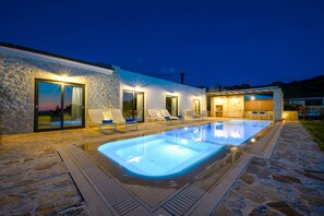 Ein moderner Poolbereich im Freien mit Steinböden, Liegestühlen und einem gut beleuchteten Gebäude mit großen Fenstern.