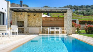 A swimming pool with a stone wall and a wooden deck, surrounded by a garden with flowers and a view of a hilly landscape. - Lithari Tria in Agios Georgios (Corfu)