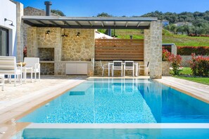 Ein Schwimmbecken mit einer Steinmauer und einer Holzterrasse, umgeben von einem Blumengarten mit Blick auf eine hügelige Landschaft.