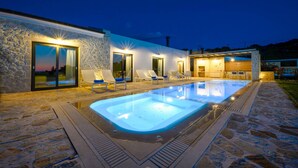 A modern outdoor pool area with stone flooring, lounge chairs, and a well-lit building with large windows. - Lithari Tria in Agios Georgios (Corfu)