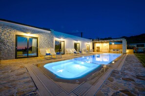 Ein moderner Poolbereich im Freien mit Steinböden, Liegestühlen und einem gut beleuchteten Gebäude mit großen Fenstern.