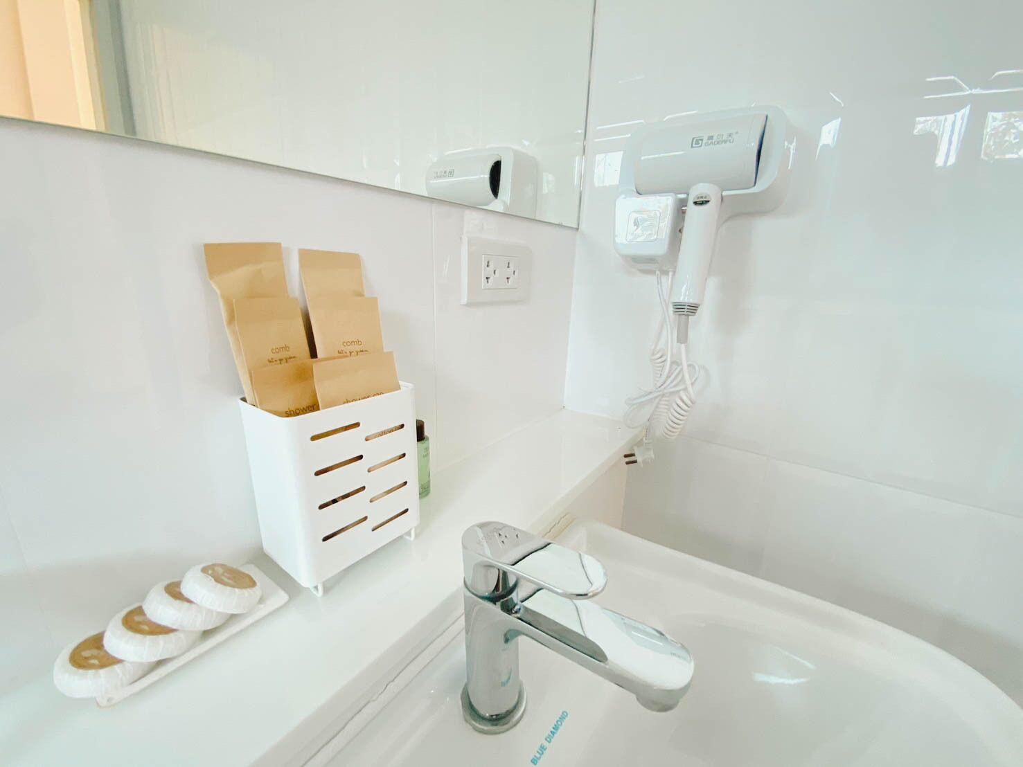 A modern bathroom with a white sink, a chrome faucet, and a hairdryer mounted on the wall. There are brown paper bags hanging on a rack and a green bottle on the counter.