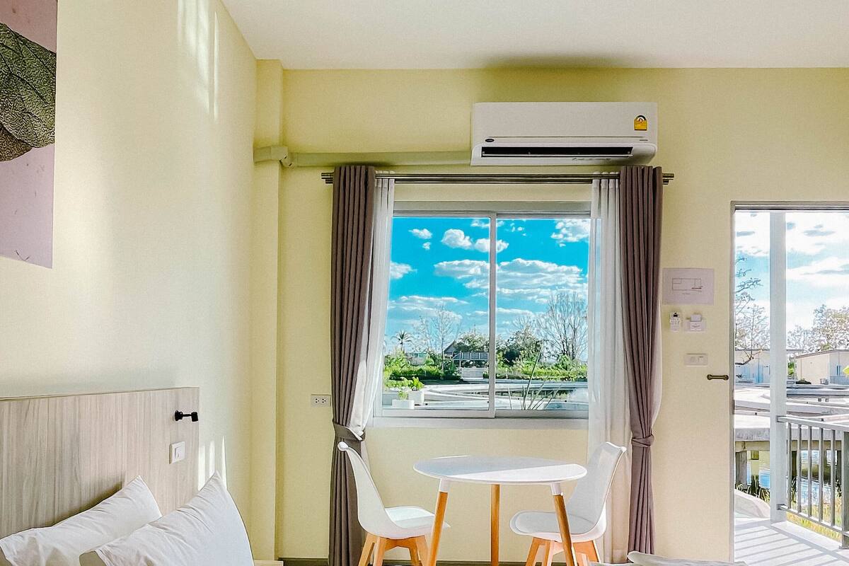 Quarto de hotel com uma cama, uma pequena mesa com cadeiras, uma janela com cortinas e um aparelho de ar condicionado.