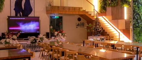 A restaurant with a long table, chairs, a TV screen, and a staircase.