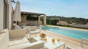 Un espace aménagé au bord de la piscine, avec des meubles blancs, une table garnie de fruits et de boissons, et une vue sur la piscine et les environs.