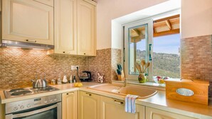 A kitchen with a brick backsplash, a stove, a sink, and a window with a view of the hills. - Villa Melissa Paxos in Paxi (Paxos)