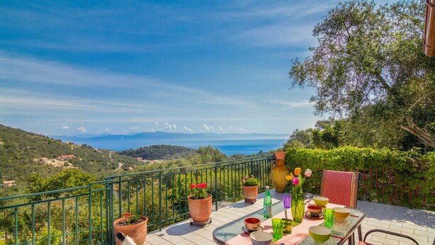 A terrace with a table set for dining, potted plants, and a view of the sea and hills. - Villa Melissa Paxos in Paxi (Paxos)