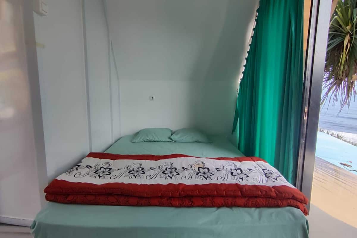 Una cama con colcha verde y manta roja y blanca, cortinas verdes y vista a la playa.