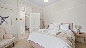 A bedroom with a bed, bedside tables, a chair, and a door leading to another room. - Hamptons House | Scandi Heritage Charm (Bathurst)