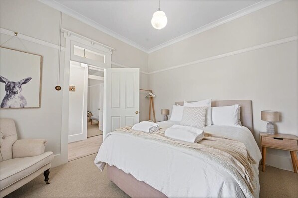A bedroom with a bed, bedside tables, a chair, and a door leading to another room. - Hamptons House | Scandi Heritage Charm (Bathurst)
