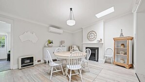 A dining area with a round table, white chairs, a chandelier, a fireplace, and a bookshelf. - Hamptons House | Scandi Heritage Charm (Bathurst)