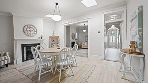 A dining area with a round table, white chairs, a fireplace, and a clock on the wall. - Hamptons House | Scandi Heritage Charm (Bathurst)
