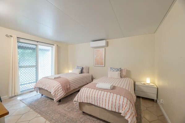 Ein Schlafzimmer mit zwei Betten, einem Fenster mit Vorhängen, einem Nachttisch mit Lampe und einer Deckenklimaanlage.