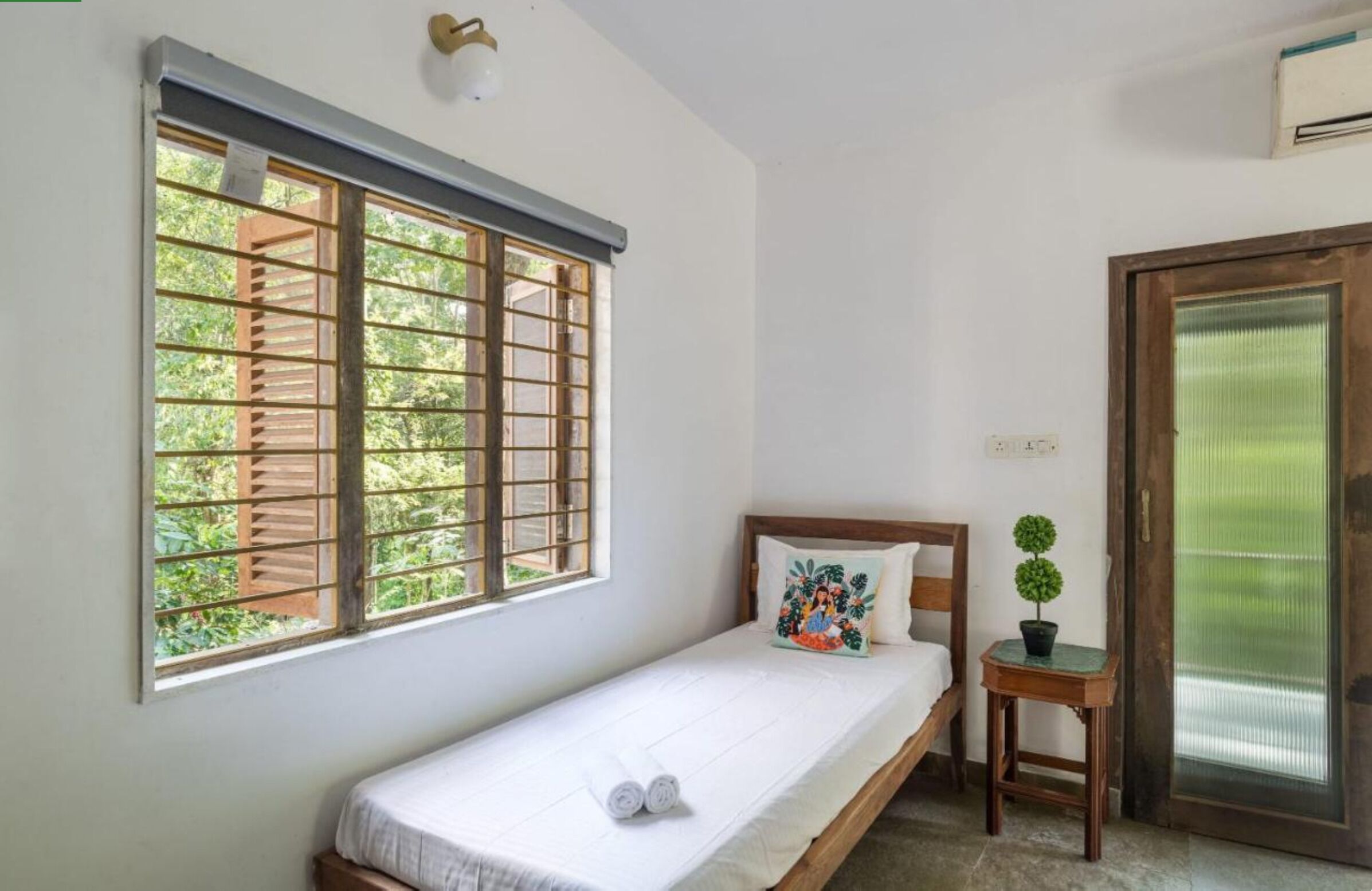Una cama individual con ropa de cama blanca, una mesita de noche pequeña con una planta y una ventana con vistas a zonas verdes.