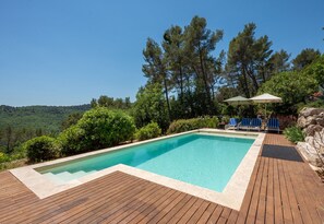 A rectangular swimming pool with a wooden deck, surrounded by greenery and trees, with a clear view of the landscape. - Mas des Violettes – Luxury vacation villa in the Alpes-Maritimes (Tourretes sur Loup)