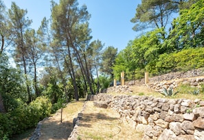 A stone pathway leading through a forested area with trees and a stone wall on one side. - Mas des Violettes – Luxury vacation villa in the Alpes-Maritimes (Tourretes sur Loup)