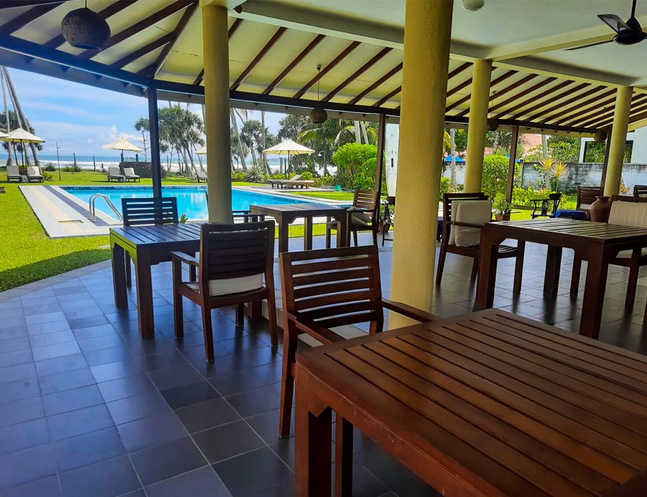 Uma área de pátio ao ar livre com mesas e cadeiras de madeira, uma piscina e vista para o oceano.